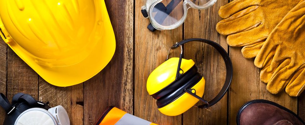 Personal safety workwear items shot directly above. The safety equipment includes a hard hat, goggles, gloves, steel toe shoes, earmuff a respiratory mask and a safety vest. The background is a rustic wood table scratched and stained with vertical stripes.  Predominant colors are yellow and brown. DSRL studio photo taken with Canon EOS 5D Mk II
